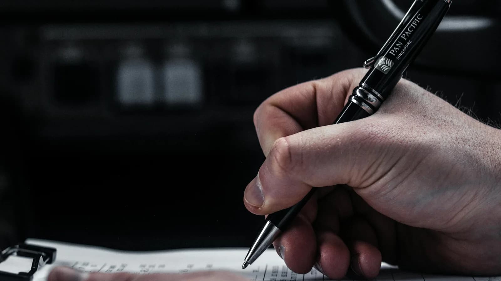 Pilot writing on a kneeboard checklist in a dim cockpit with glass-panel avionics behind — pre-flight paperwork in operator context