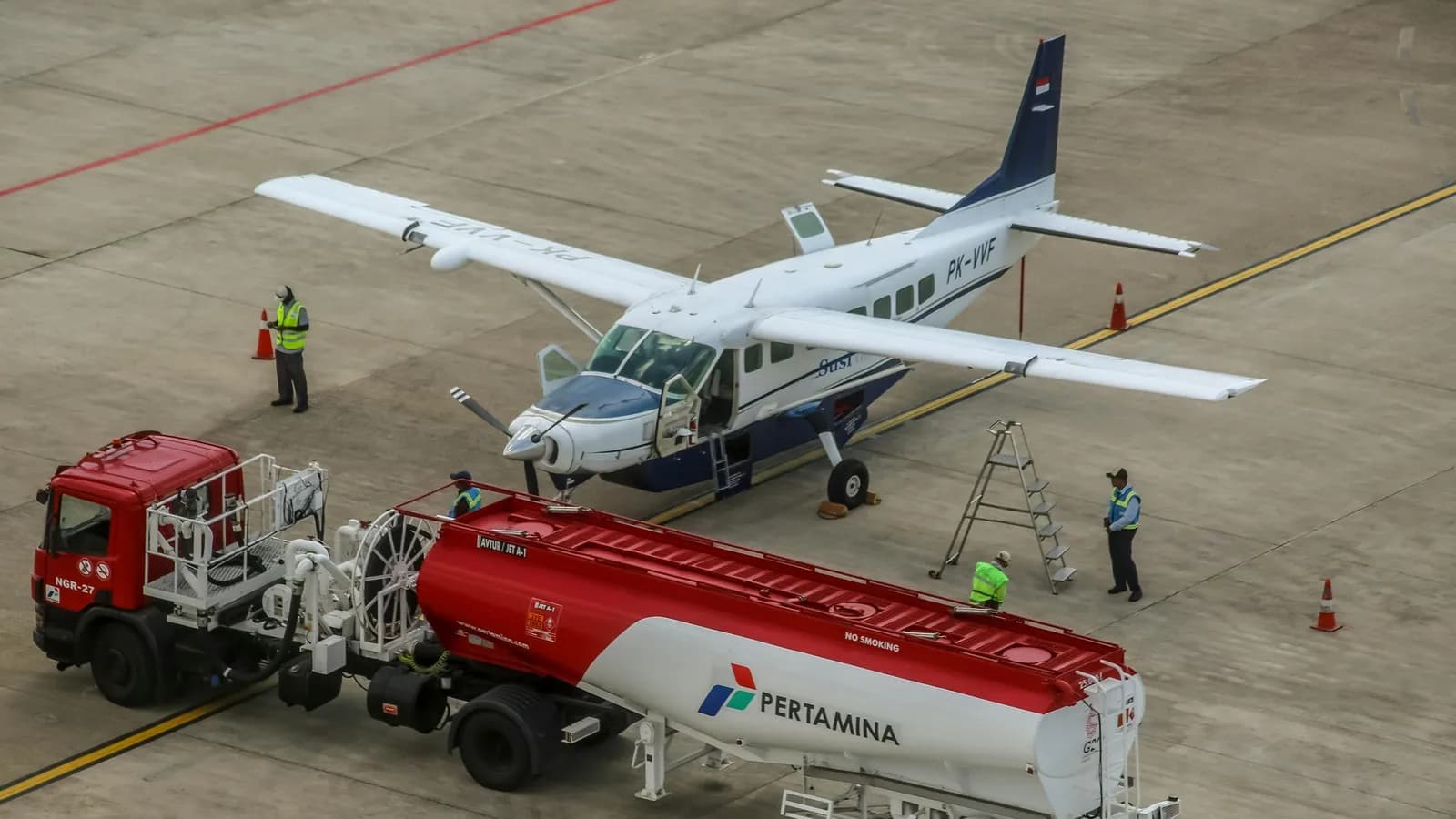 Ground crew refuelling a Cessna Caravan from a Jet A-1 bowser on an independent FBO ramp, with chocks, access ladder and safety cones in place