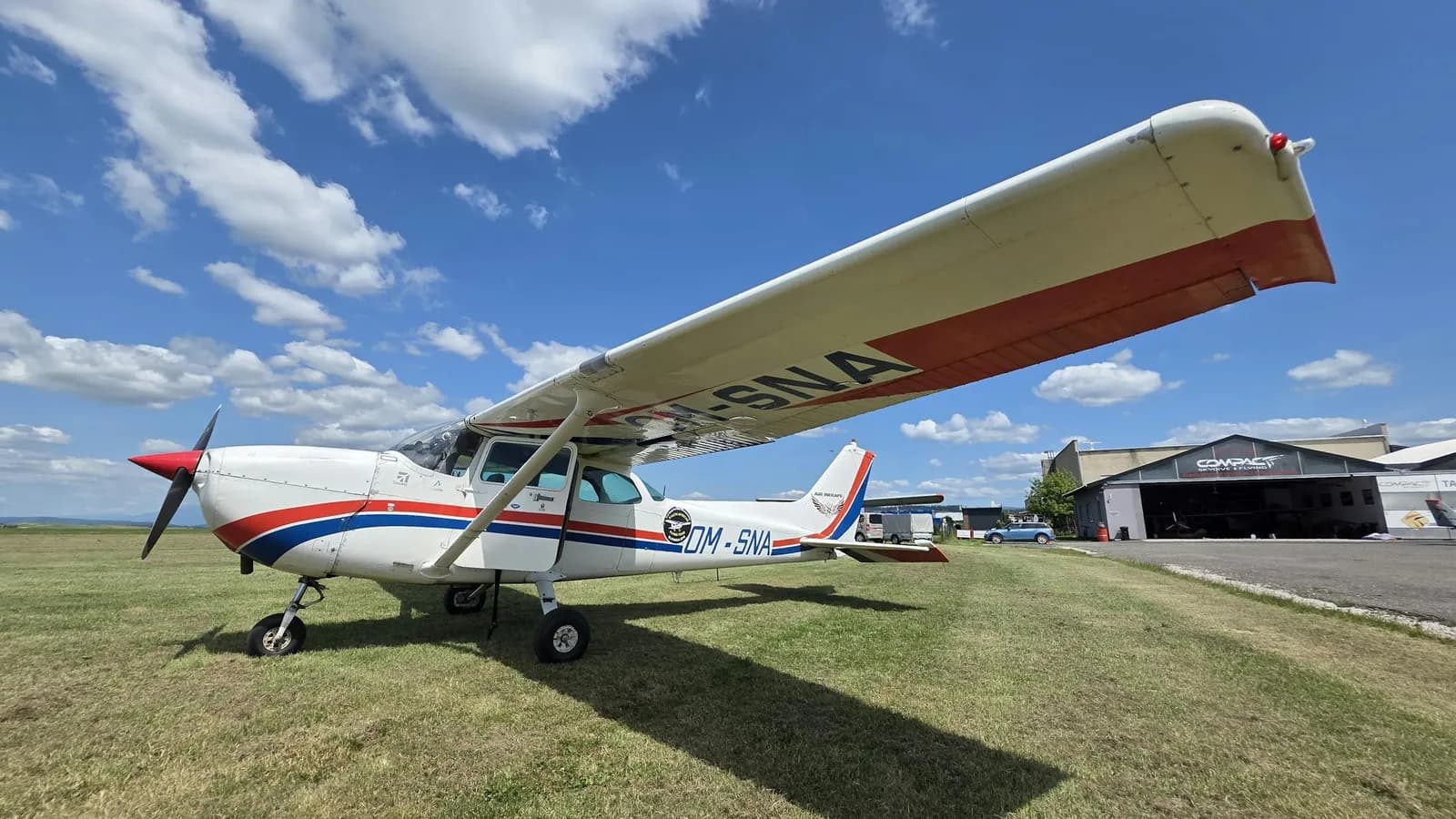 Cessna 172 single-engine training aircraft parked on a grass apron with a hangar behind — flight school ramp scene